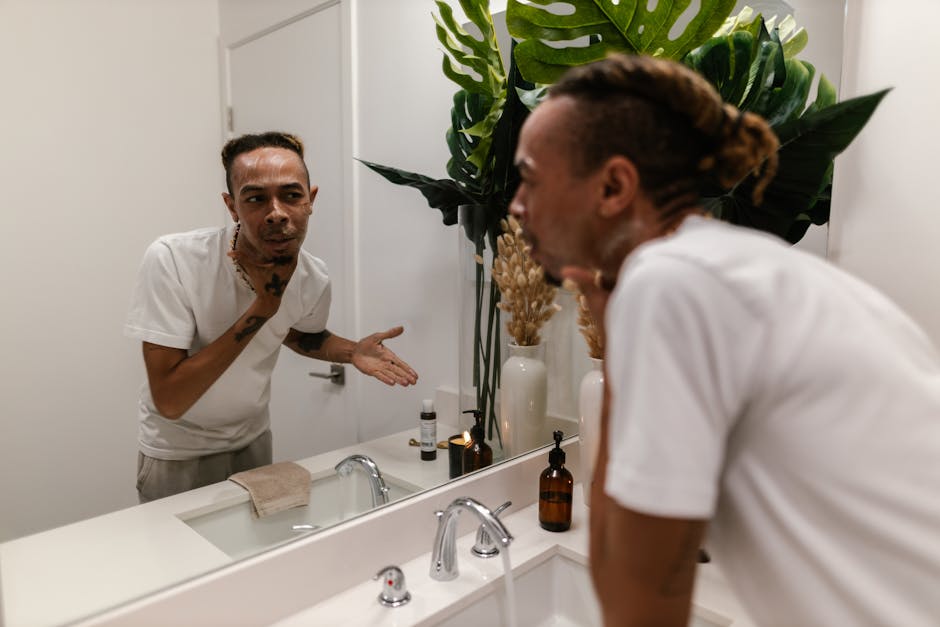 A young man applying skincare product while looking in the mirror in a modern bathroom.