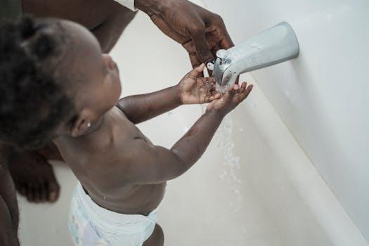 A toddler being guided by an adult to wash hands under a faucet, promoting hygiene.