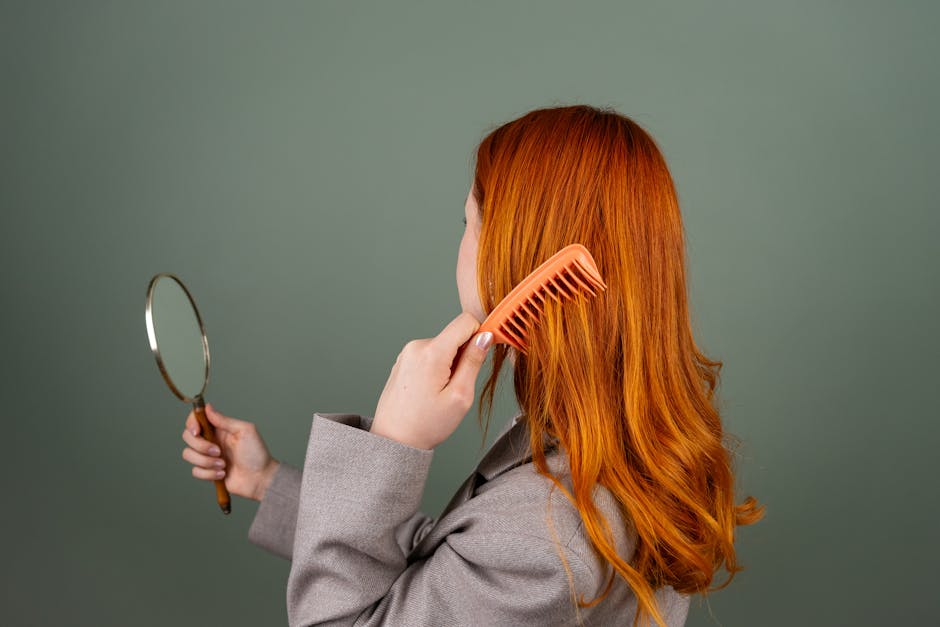 Red-haired woman using a comb while holding a mirror, focusing on hair care and beauty.