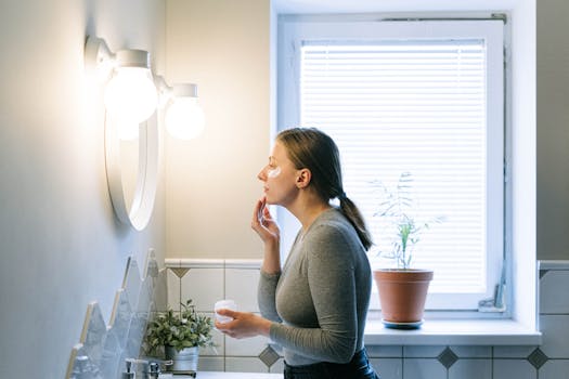 A woman applies face cream in a well-lit bathroom, emphasizing skincare routine.