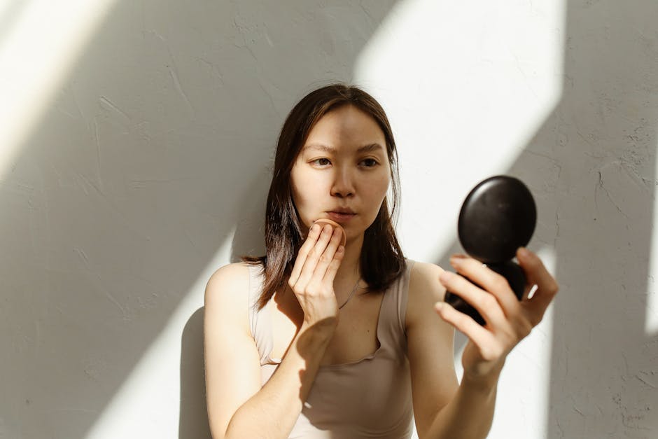 Young Asian woman applying makeup with a mirror in soft natural lighting indoors.