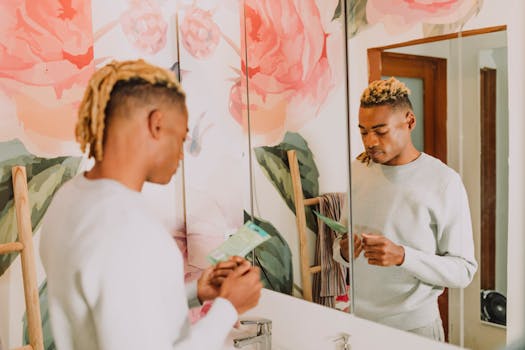 A man stands in a bathroom with floral decor, holding a skincare product and reflecting in the mirror.